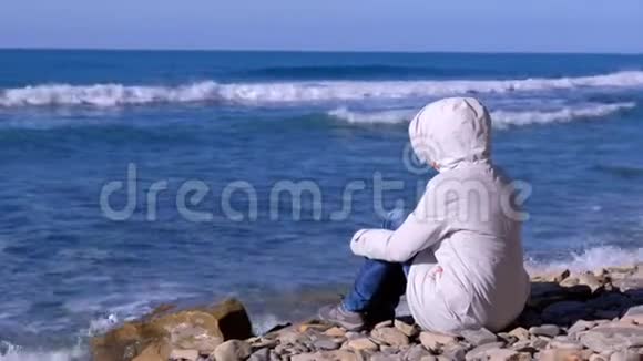 穿着白色羽绒服和风帽的女人坐在岸边看着汹涌的海浪拍打着石头视频的预览图