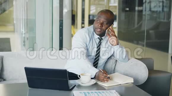 一位非洲裔美国商人在智能手机上聊天在他坐着的笔记本上写下商业信息视频的预览图