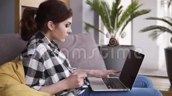 放松的漂亮女孩舒适地坐在沙发上笔记本电脑在她的腿上手里拿着一张卡片正在打字视频的预览图