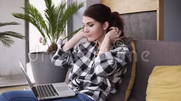 漂亮的年轻女人坐在沙发上放松戴上耳机打开笔记本电脑上的音乐微笑着走向视频的预览图