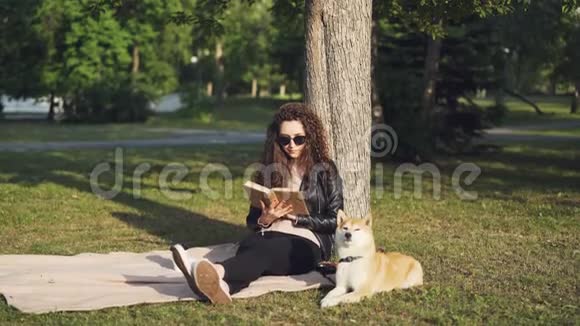 有魅力的女学生正在看书坐在城市公园树下的格子上小狗躺在附近享受着快乐视频的预览图