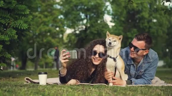 年轻女子正在公园里和男朋友和宠物狗躺在草地上自拍可爱的动物在打喷嚏和视频的预览图
