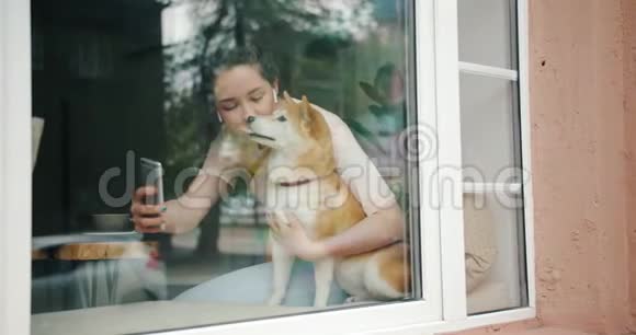 女孩自拍与狗坐在窗台在咖啡馆使用智能手机视频的预览图