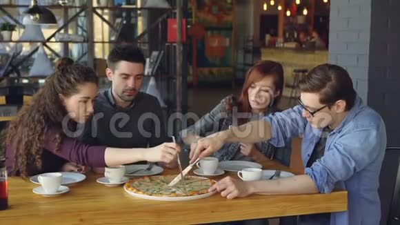 快乐的年轻人正在用刀子切大披萨在舒适的咖啡馆里吃饭时谈笑风生现代生活方式视频的预览图