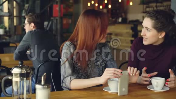 两个穿着休闲服的漂亮女人正在咖啡馆里和女孩聊天拿着咖啡杯和智能手机不错视频的预览图
