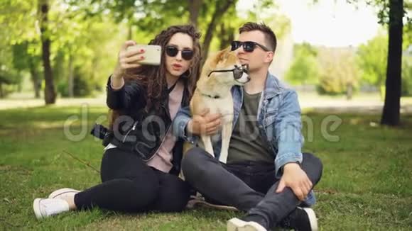 漂亮的女人正在和男朋友和可爱的狗在公园里休息时用智能手机自拍视频的预览图