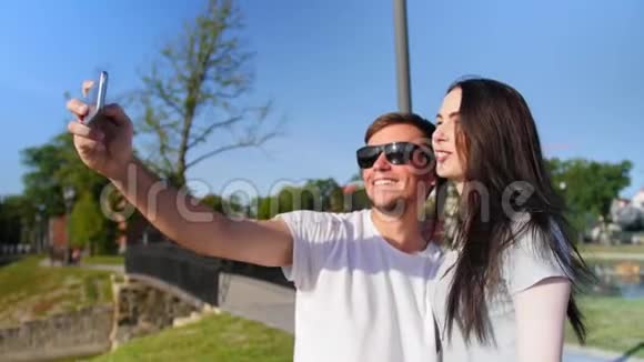 中镜头的快乐年轻夫妇自拍与智能手机同时站在公园附近的池塘中弹视频的预览图