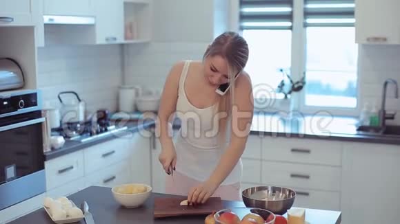 美丽的白种人女孩穿着白色衬衫在轻盈的厨房里做饭在手机上说话微笑着视频的预览图