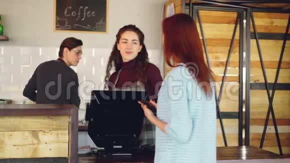 开朗的女性收银员把外卖咖啡卖给年轻女性并接受智能手机付款但很好视频的预览图