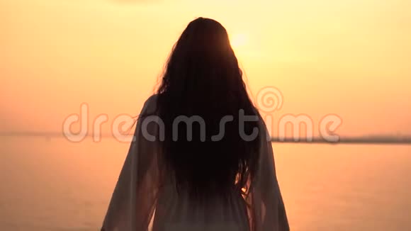 一个穿着海滩礼服的女孩站在夕阳背景上双手朝不同的方向伸展慢动作高清高清视频的预览图