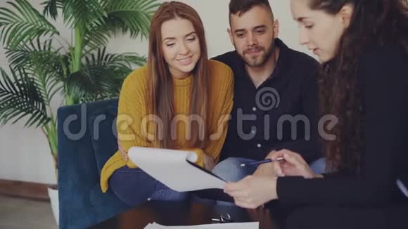 可爱的一对夫妇正在与房地产经纪人在漂亮的新公寓见面时检查文件年轻人坐在沙发上视频的预览图