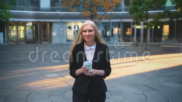 在院子里喝咖啡的正式女人优雅的金发女郎穿着西装留着长发喝着白色杯子里的咖啡视频的预览图