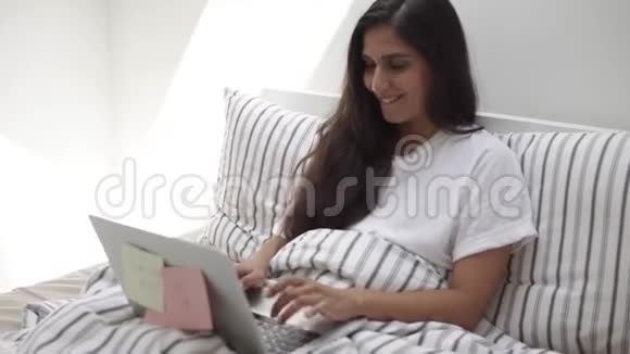 微笑的黑发女人坐在床上靠在枕头上在笔记本上打字视频的预览图