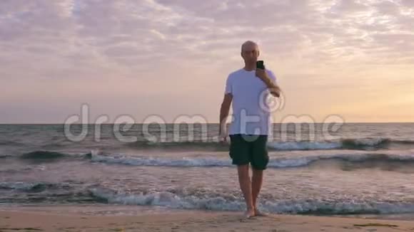 日出时在波浪海附近的海滩上男人在智能手机上自拍视频的预览图