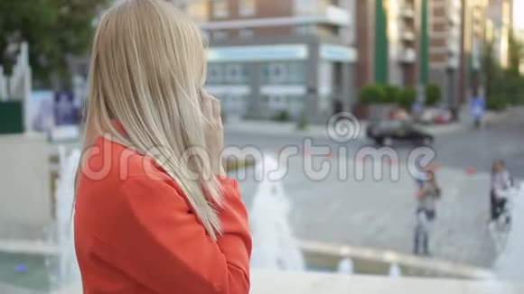 年轻漂亮的金发女郎穿着红色夹克在喷泉背景下打电话视频的预览图