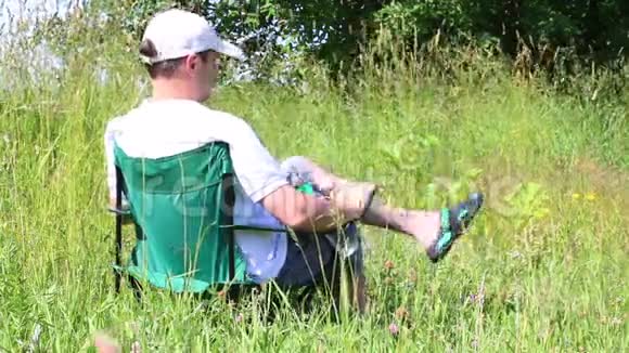 一个男人坐在一个有灯光的草地上的野餐椅上休息时他双手合拢在胸前药草和花从夏天摇曳而出视频的预览图