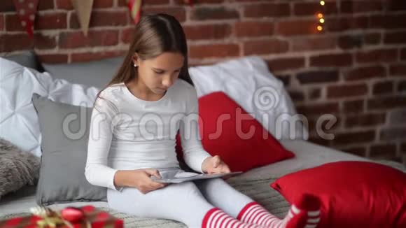 圣诞快乐的小女孩帕贾玛用圣诞老人送给她的数码平板电脑坐在卧室里装饰着家视频的预览图