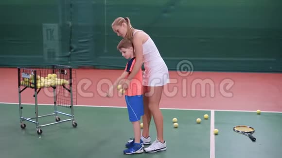 身着白色短裙的女教练正在教小男孩在室内球场玩耍职业网球视频的预览图