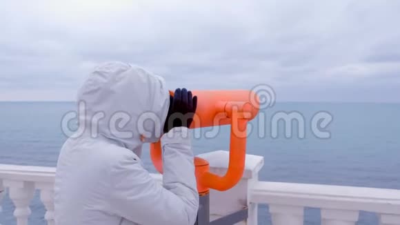 穿着白色夹克和引擎盖的女人用望远镜在海边看海景侧视视频的预览图