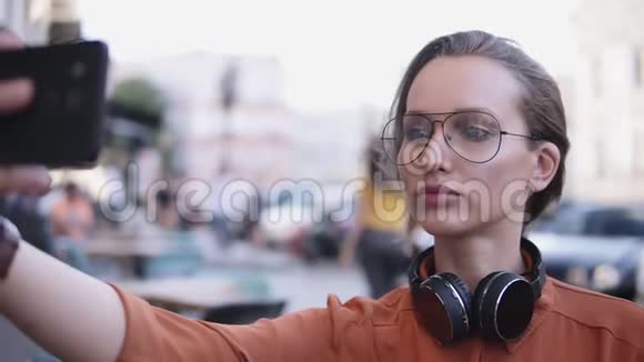 年轻的女人白天化妆站在街上用她的智能手机自拍模糊的街道视频的预览图