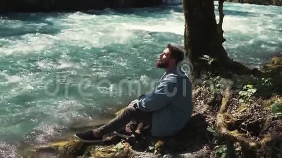 一个留着胡须的年轻人坐在一棵孤独的树和一条河旁边休息视频的预览图