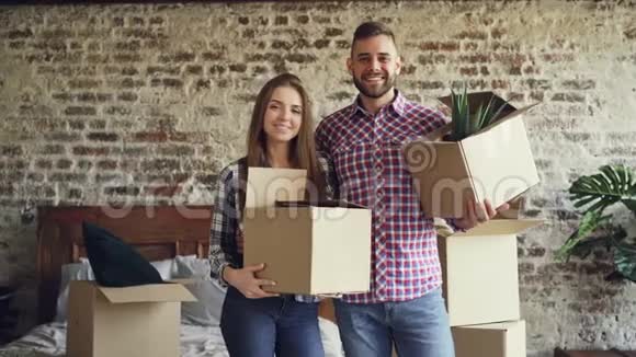 幸福的年轻夫妇站在新房子里拿着纸箱微笑着看着相机的肖像家庭视频的预览图