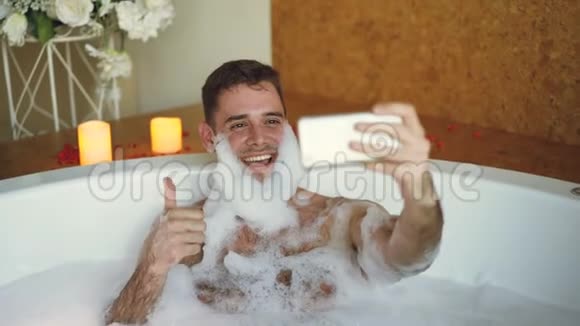 帅哥正在浴缸里用智能手机拍自拍胡子上有肥皂泡沫他在笑在做视频的预览图