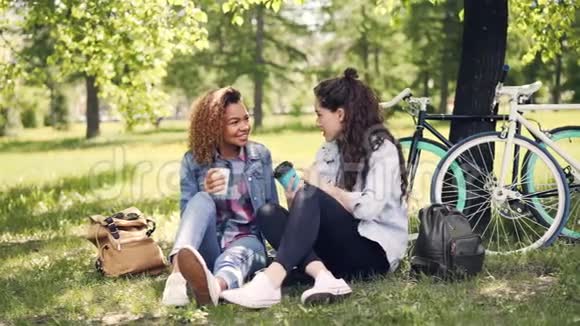 快乐的女孩在外面聊天喝着外卖咖啡坐在公园的草坪上背包和自行车在里面视频的预览图
