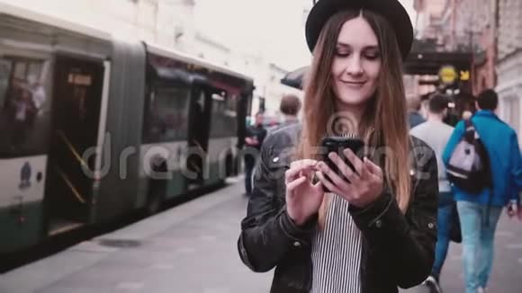 美丽的年轻快乐的欧洲女孩戴着时尚的帽子在繁忙的城市街道上行走使用智能手机应用程序慢动作视频的预览图