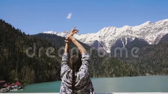 一位妇女双手举着站在蓝湖和白雪皑皑的山前视频的预览图