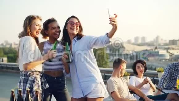 漂亮的年轻女士在屋顶的聚会上用智能手机装瓶子自拍而她们的朋友则是这样视频的预览图
