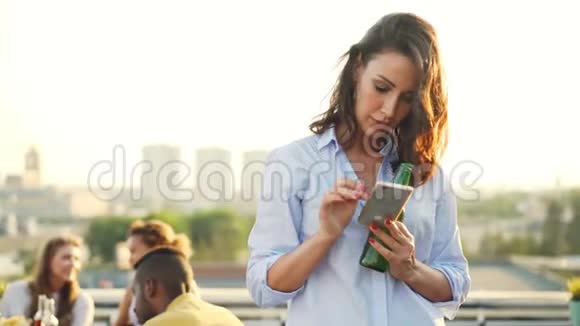 拉拉队的黑发美女使用智能手机触摸屏幕站在屋顶手里拿着啤酒瓶视频的预览图
