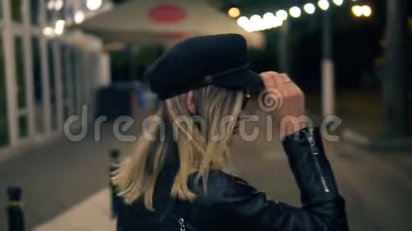一个漂亮的女孩戴着时髦的帽子穿着黑色的皮夹克在一盏咖啡馆的灯光下穿过了夜间公园视频的预览图
