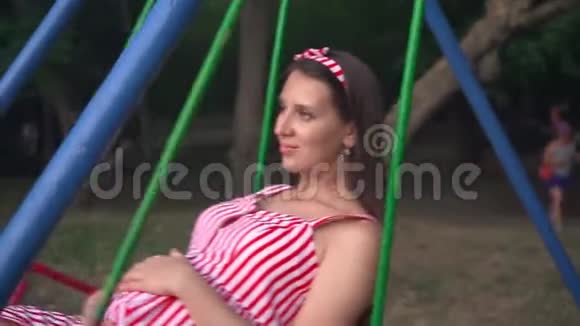 公园里的怀孕女孩在秋千上摇摆快乐的女孩一头乌黑的长发穿着条纹的白红色连衣裙在秋千上视频的预览图