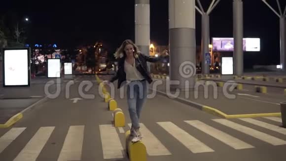 年轻的金发女人穿着牛仔裤和黑色皮夹克晚上在黄色的街道保险杠上跳跃玩得很开心街道视频的预览图