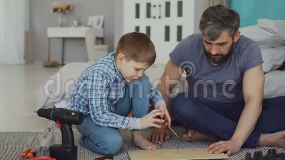 严肃的小男孩专心致志地在父亲帮助他的时候用螺丝刀把木头碎片放好视频的预览图