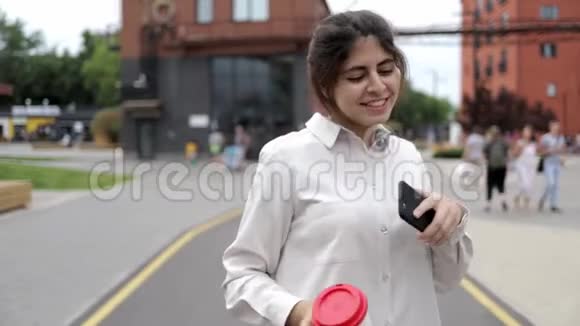 有魅力的年轻女孩带着咖啡和智能手机在街上跳舞视频的预览图