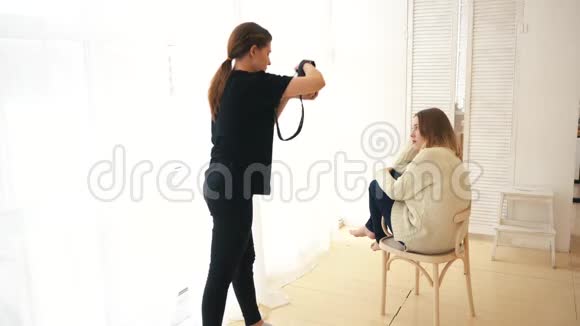 年轻的女摄影师在坐在木制的白色套头衫和深色牛仔裤上拍摄微笑模特视频的预览图