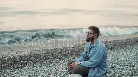 一个年轻有胡子的男人坐在石滩上欣赏海景视频的预览图