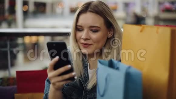 一个漂亮的金发女孩在拿着购物袋坐在咖啡馆里的手机视频的预览图