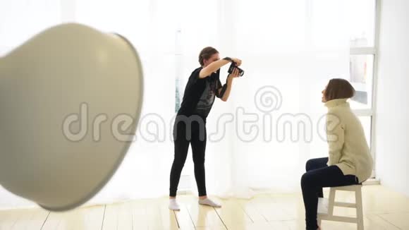 年轻的女摄影师在坐在木制的白色套头衫和深色牛仔裤上拍摄微笑模特视频的预览图