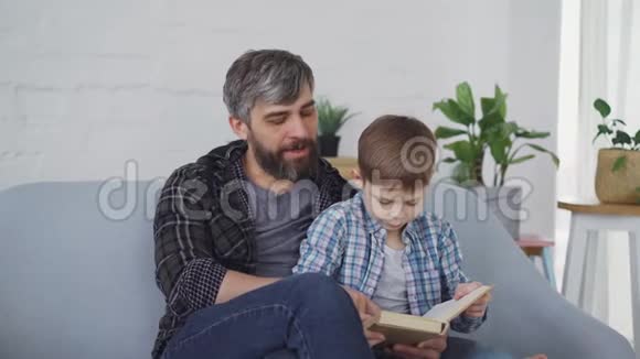 关爱的父亲正在教他的小儿子好奇的学龄前儿童阅读他们坐在沙发上看书视频的预览图