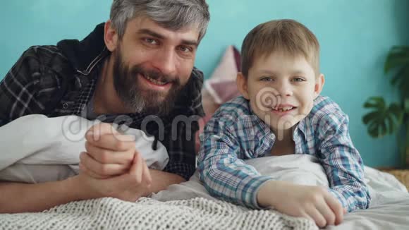 特写肖像两人成年父亲和可爱的小儿子躺在家里的床上微笑父爱视频的预览图
