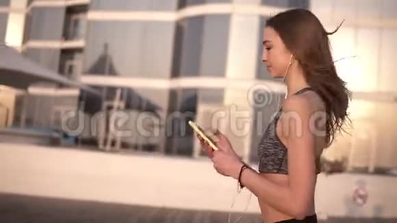 年轻女子在户外跑步女健身女孩在室外进行慢跑训练用耳机听音乐视频的预览图