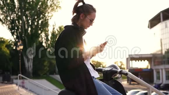 穿着黑色连帽衫的年轻女孩坐在摩托车上在手机上打字坐在公园外面的摩托上太阳视频的预览图