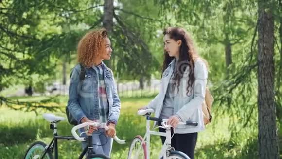 漂亮的女孩现代学生在公园里聊天谈笑风生地拿着绿色漂亮的自行车视频的预览图