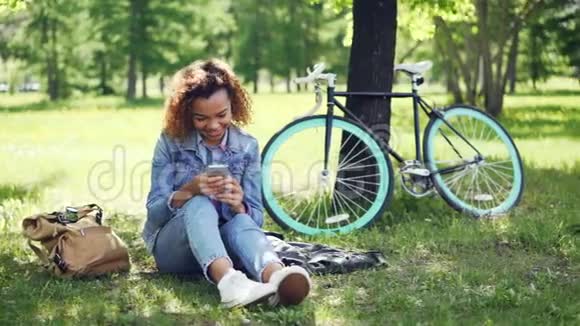 快乐的非裔美国女孩骑自行车后在公园休息时使用智能手机触摸屏幕和微笑视频的预览图