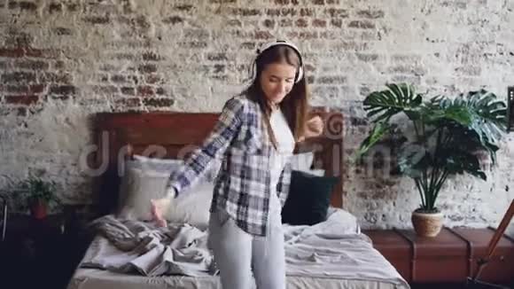 穿着休闲服的漂亮粗心的女孩在卧室里跳舞用无线耳机听音乐青年视频的预览图