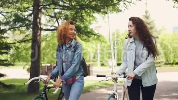 快乐的游客美丽的女人带着自行车和背包在美丽的绿色公园里散步聊天自行车视频的预览图