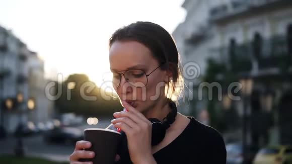 年轻女子戴着时尚太阳镜在街上喝着一杯带塑料吸管的咖啡傍晚时分太阳照耀着视频的预览图
